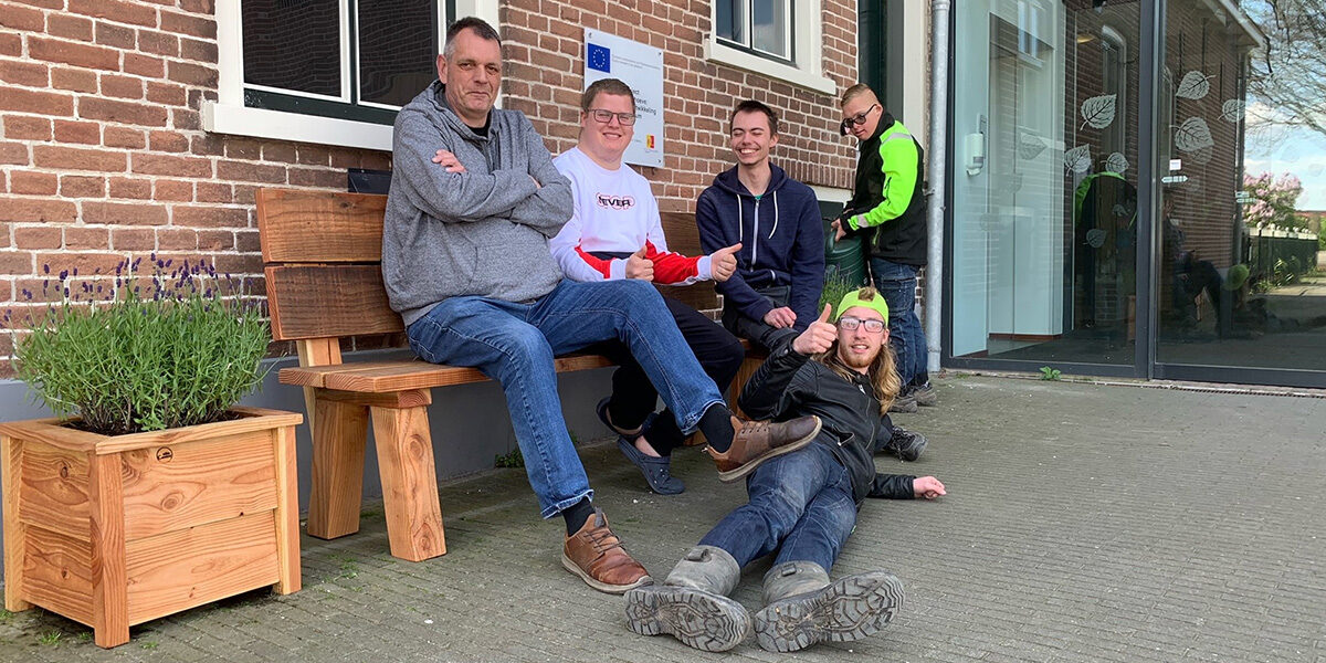 een groep mannen zit, ligt en staat op en rond een houten tuinbank